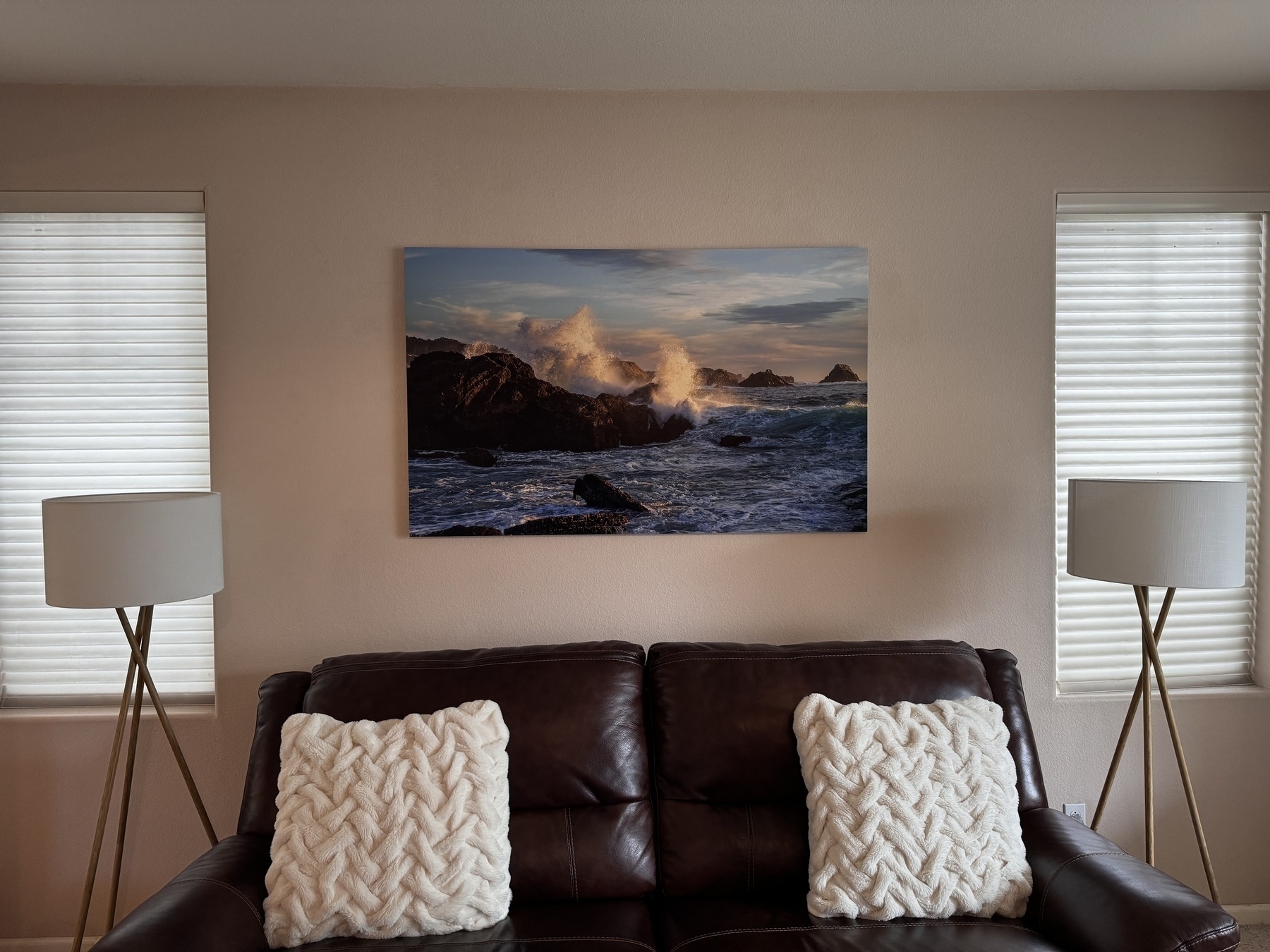 Photo of a living room, with a couch, two lamps, and a large photograph of a wave crashing onto rocks mounted onto the wall behind the couch.